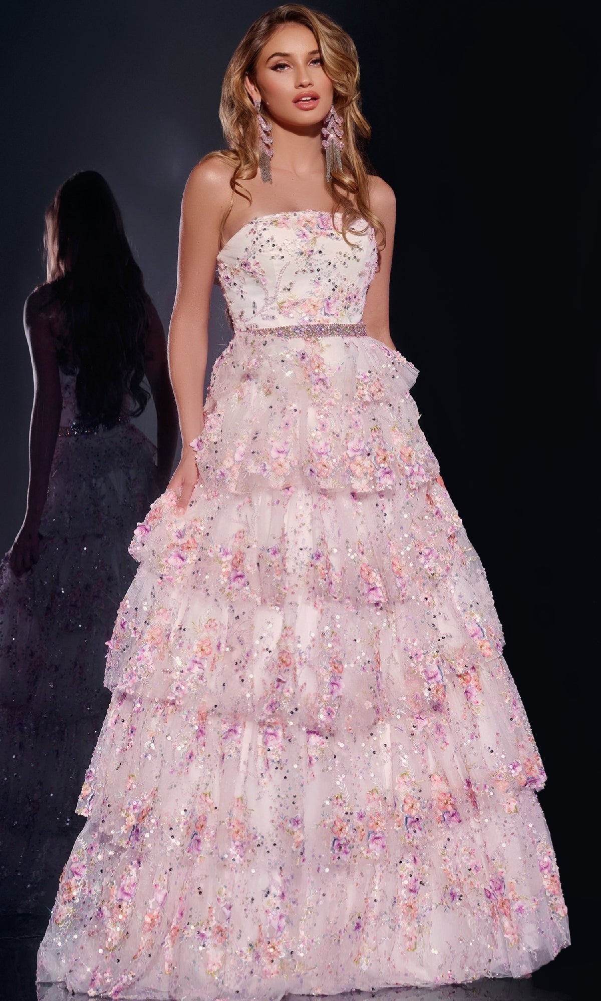A woman wears the Jovani 42190 Long Prom Dress—a strapless gown with a tiered skirt, beaded waistline, and pastel floral lace with shimmering sequins on a dark background. Her reflection appears behind her.